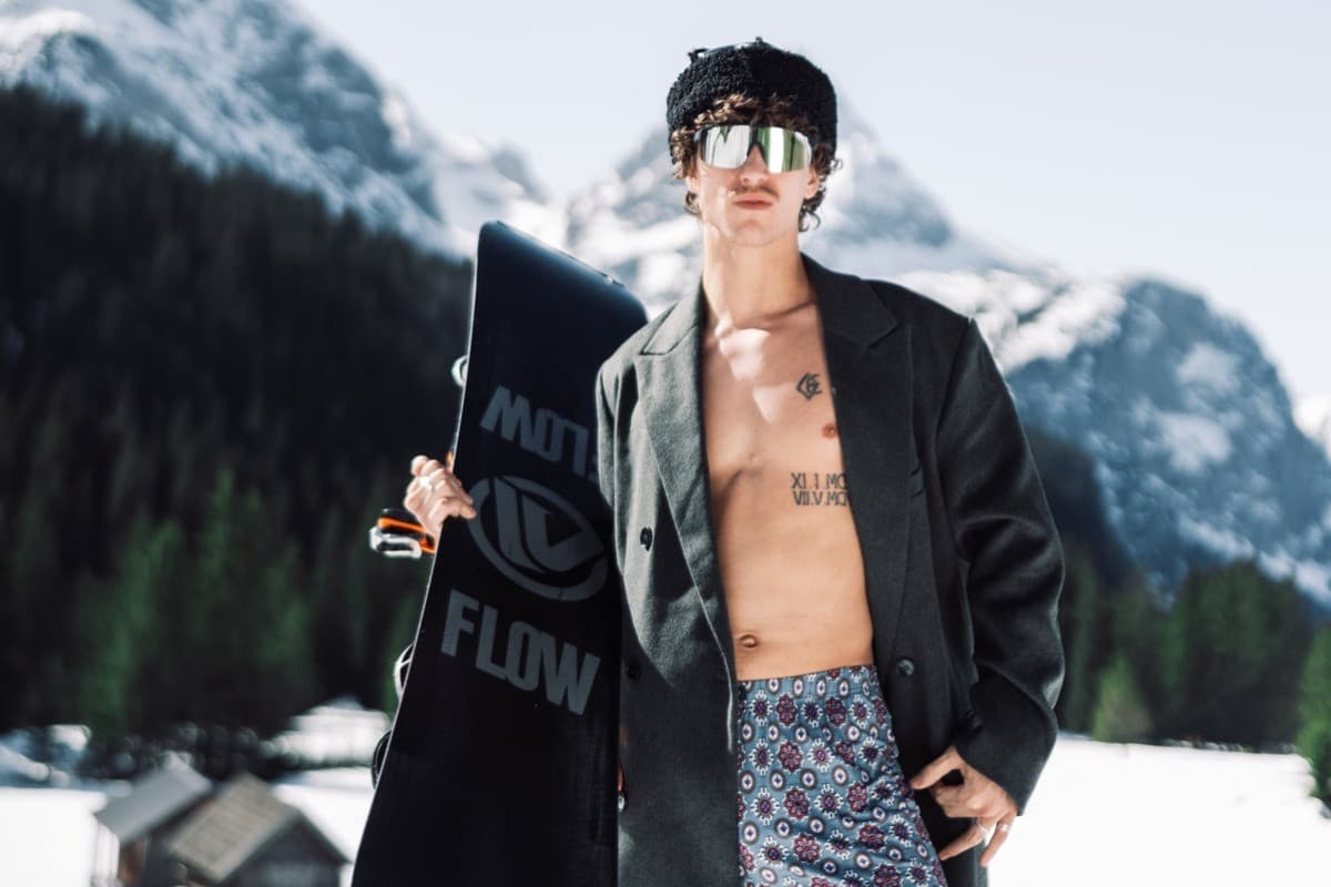 Männliches Model steht mit einem Snowboard auf dem Arm im Schnee und trägt unter einem schwarzen Mantel eine kurze, blau-gemusterte Hose. | mey®